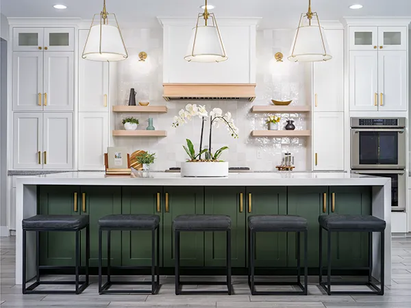 A stunning kitchen featured on Stately’s About Us page, showcasing a large island with a green base and sleek gold hardware, paired with modern black stools. The space is accented by white cabinetry, open wooden shelves, brass light fixtures, and a center orchid arrangement, embodying Stately’s luxury home staging and interior design expertise.