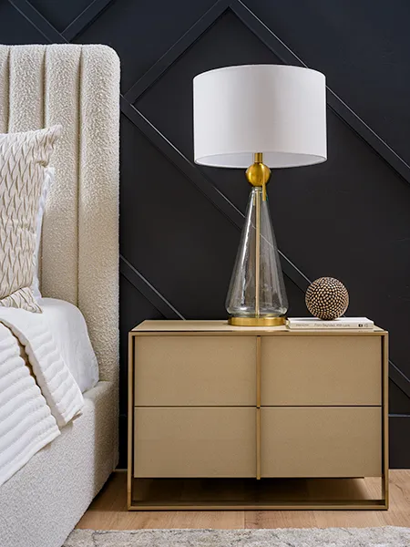 A stylish bedroom nightstand with a textured bed frame, a glass and gold lamp, and a dark accent wall with geometric detailing.